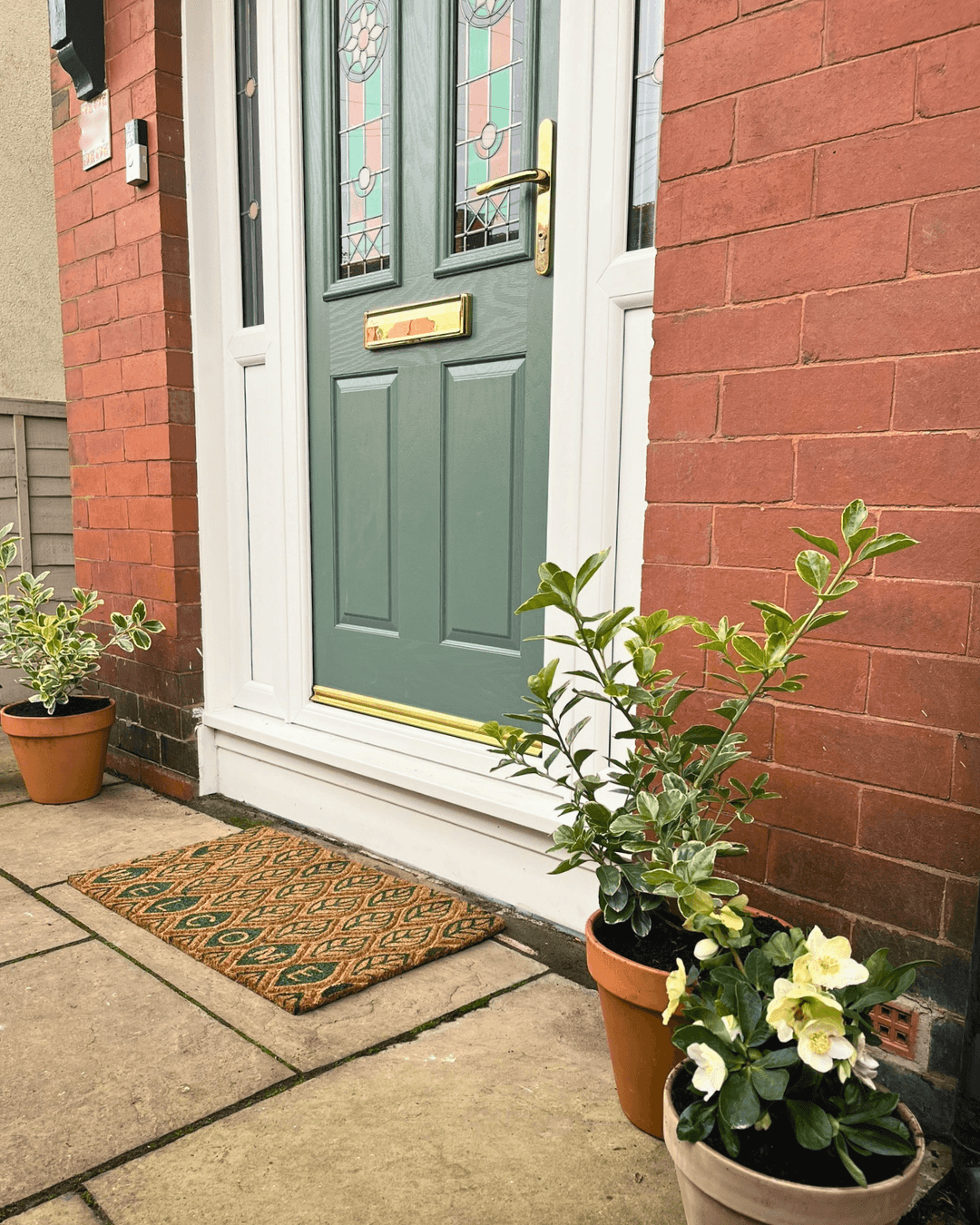 Green front door entrance styled with potted plants and patterned doormat outside red brick home